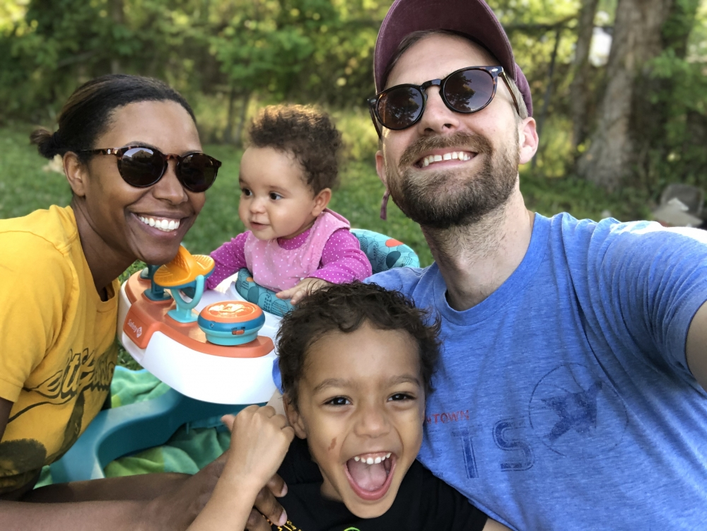 A smiling family of four enjoying a sunny day outdoors. The mother and father are wearing sunglasses, and two young children are in front of them. A baby is in a walker, while an older child is making a playful expression. Trees are in the background.