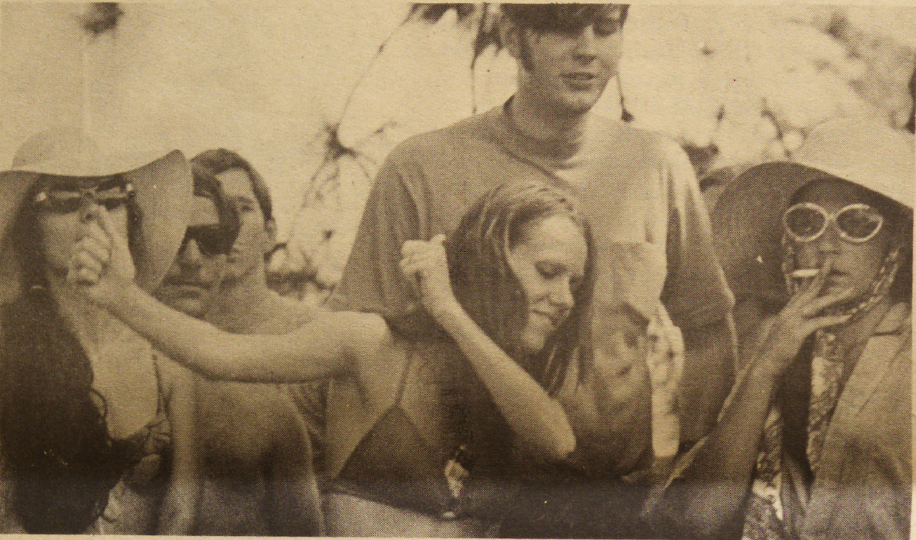 A group of people in a vintage photo, some wearing sunglasses and wide-brimmed hats. One person holds up a peace sign while another smokes. The atmosphere is relaxed and casual.