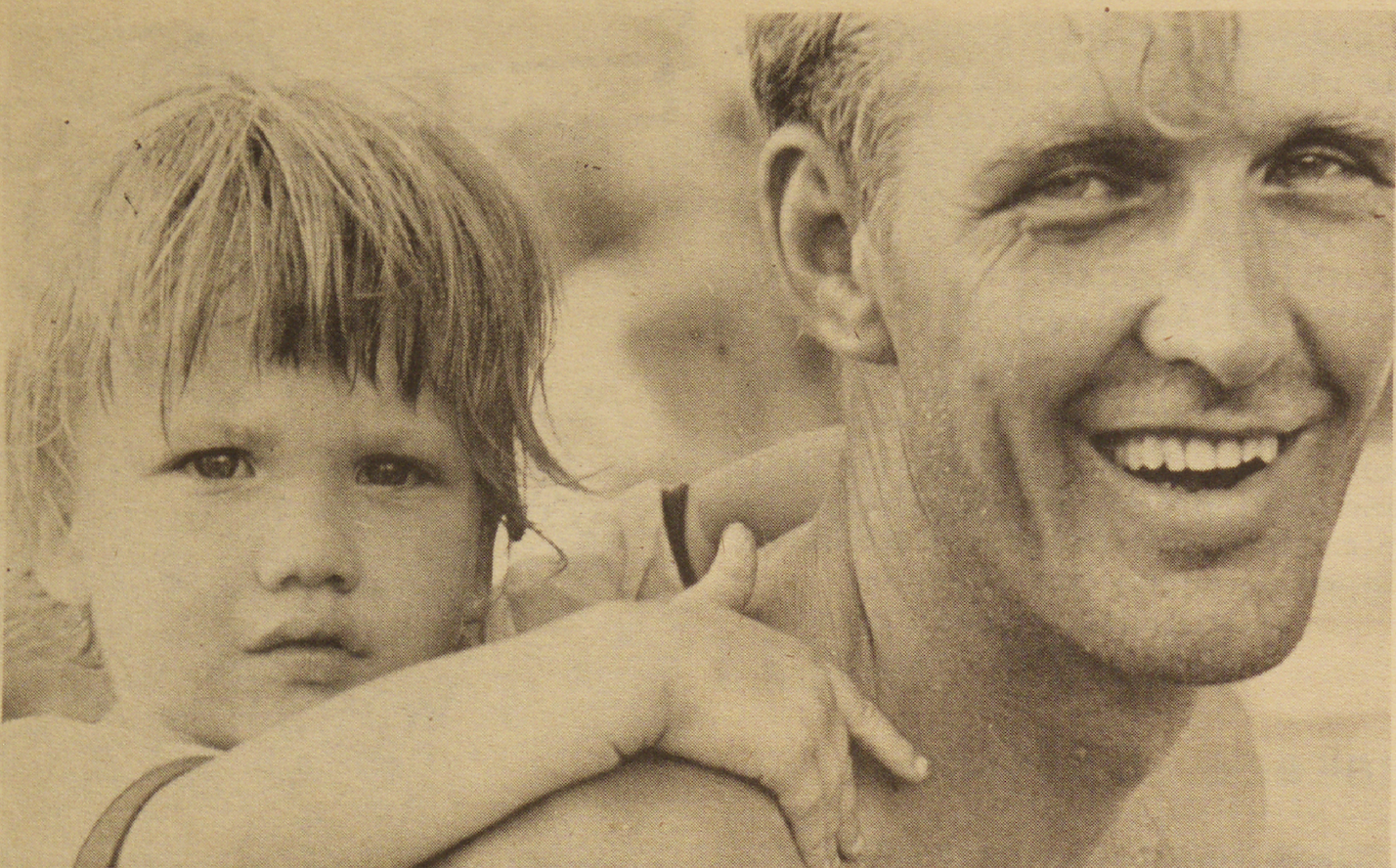 A sepia-toned photo of a smiling man with short hair carrying a young child on his back. The child, with short hair, gazes at the camera, resting their hand on the mans shoulder.