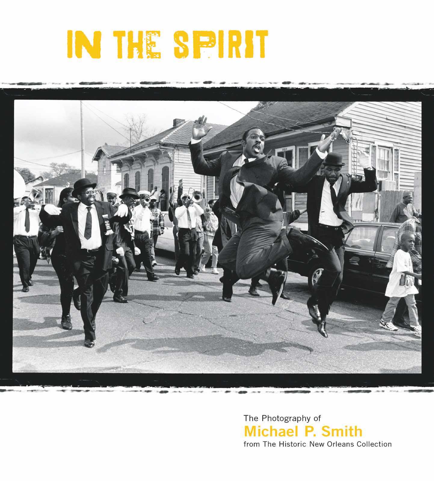 A lively black-and-white photo of a parade in New Orleans. People are dancing exuberantly in the street, dressed in formal attire with hats. The scene is vibrant with energy and joy.
