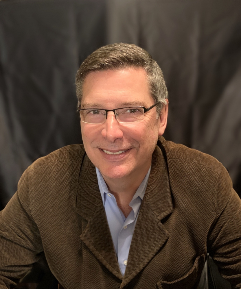 A person with short dark hair and glasses is smiling. They are wearing a brown jacket over a light blue shirt. The background is a dark, plain curtain.