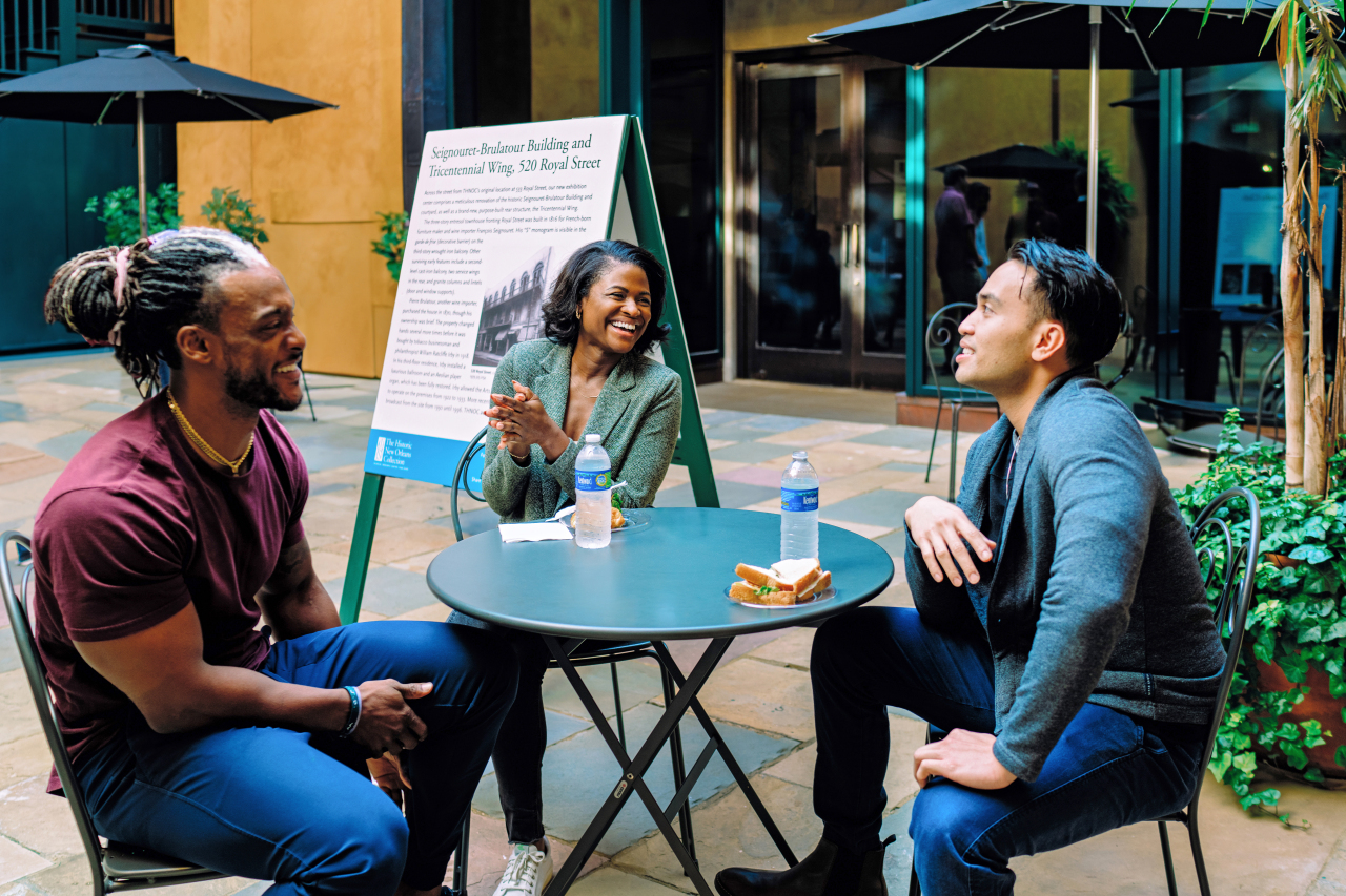 Three people sit around a small outdoor table, smiling and talking. One person holds a hot dog. A poster display is in the background along with potted plants and umbrellas.