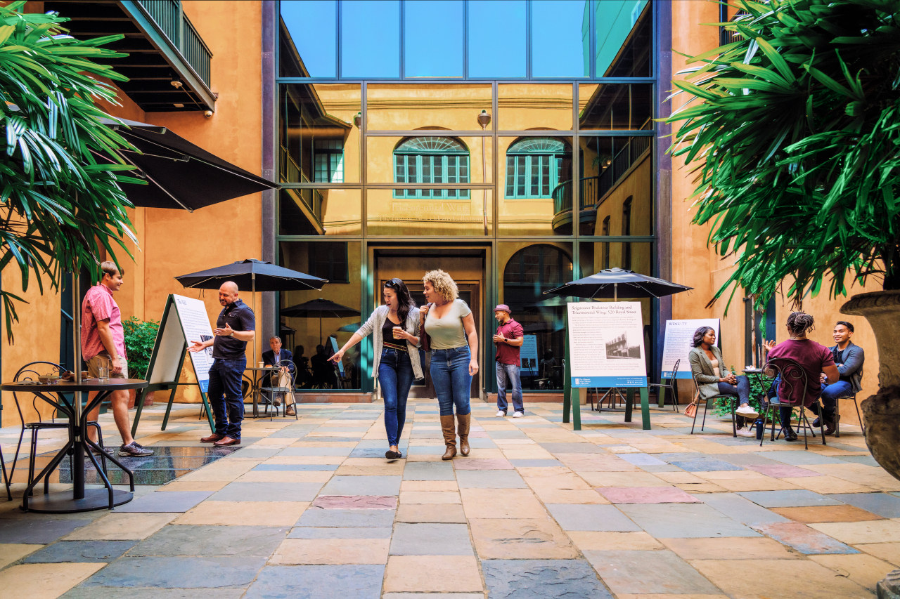 A vibrant courtyard with people walking and sitting at tables under umbrellas. The area is surrounded by tall buildings with large windows, and potted plants add greenery to the scene. Posters are displayed, and the atmosphere is lively and social.
