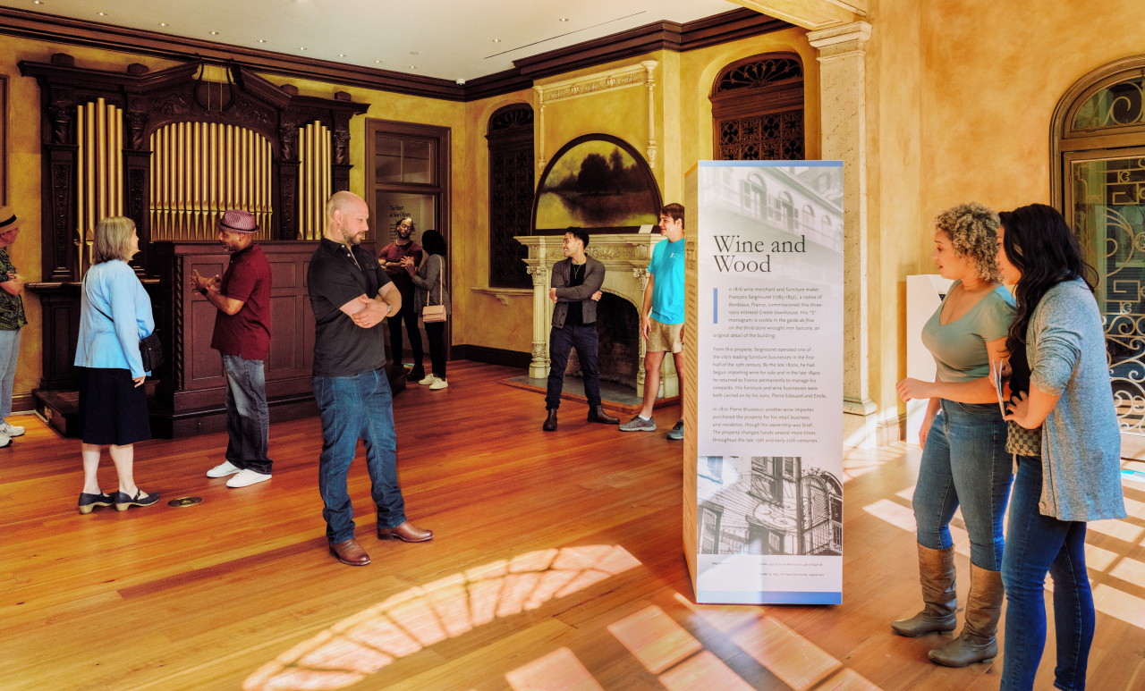 A group of people stand in a sunlit room with wooden floors, observing a display titled Wine and Wood. The room features a large organ and a fireplace. Some people are reading the display, while others look around the space.