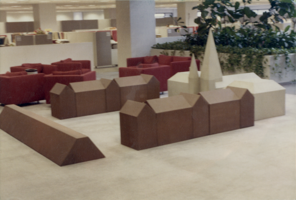 A minimalistic geometric sculpture resembling a small village is displayed on the floor of a spacious office area with red couches and large indoor plants in the background.