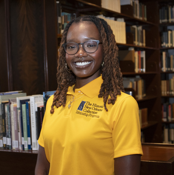 A person with glasses and long braided hair smiles while standing in a library. They are wearing a yellow polo shirt with The Historic New Orleans Collection Internship Program logo. There are shelves filled with books in the background.