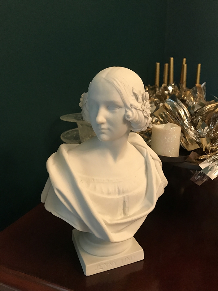 A white bust of a woman with intricate hair detail sits on a wooden surface. Behind it, there are candle holders and decorative items, set against a dark green background.