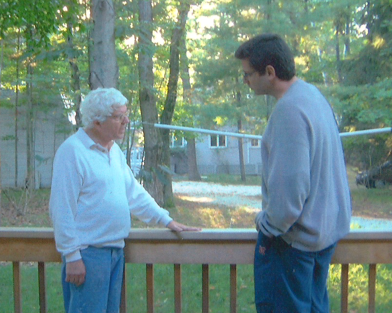 Two men stand on a wooden deck surrounded by trees. One man with white hair wears a white polo and jeans. The other man, with dark hair, wears a gray sweater and blue pants. They are engaged in conversation in a serene forest setting.