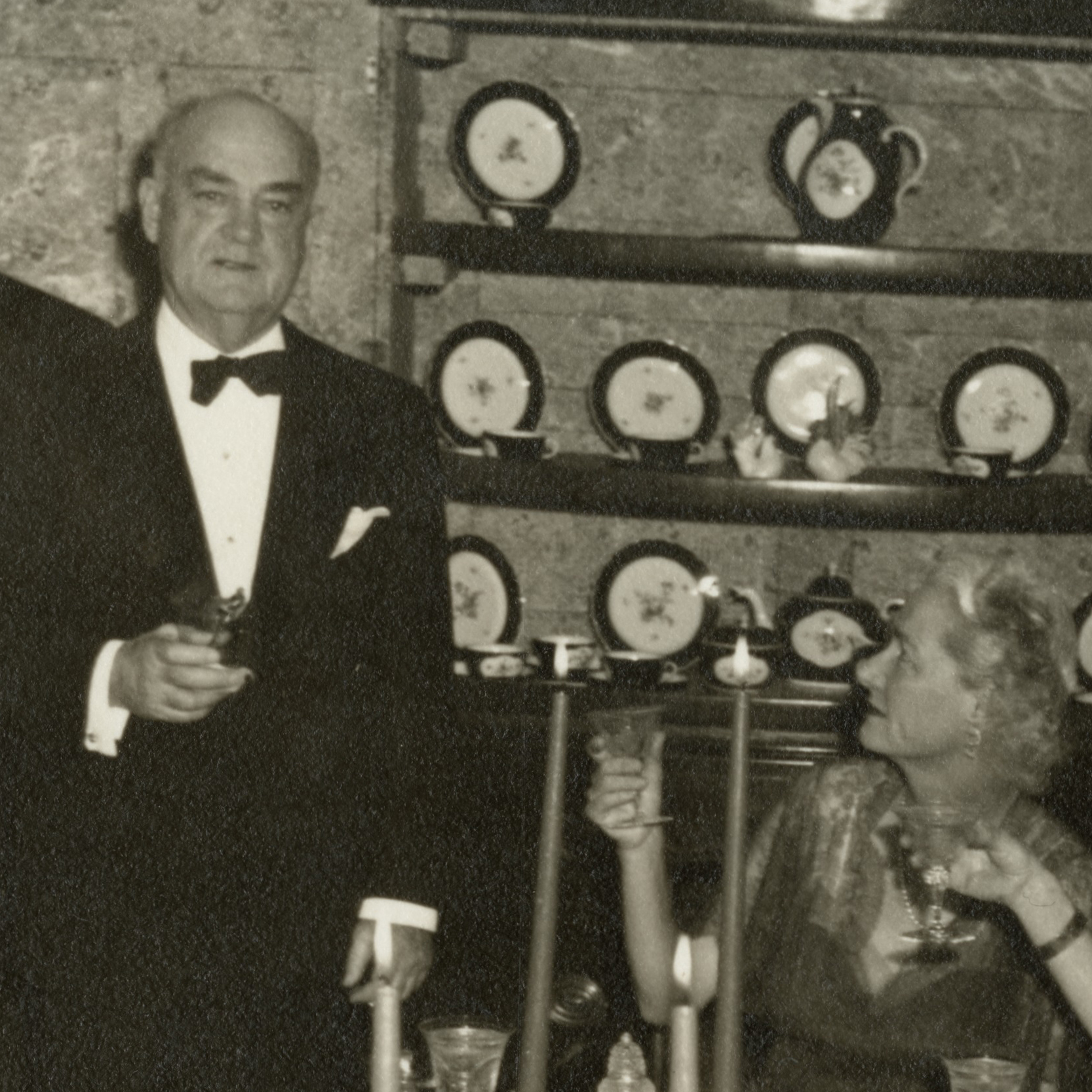 A black-and-white photo of a man in a tuxedo and a woman in a formal dress holding drinks. They are in front of a wall displaying a collection of decorative plates and teapots. The scene suggests a formal gathering or celebration.