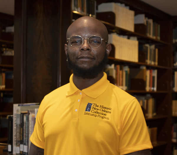 A person wearing a yellow polo shirt with the logo of The Historic New Orleans Collection Internship Program stands in a library, with shelves of books in the background.