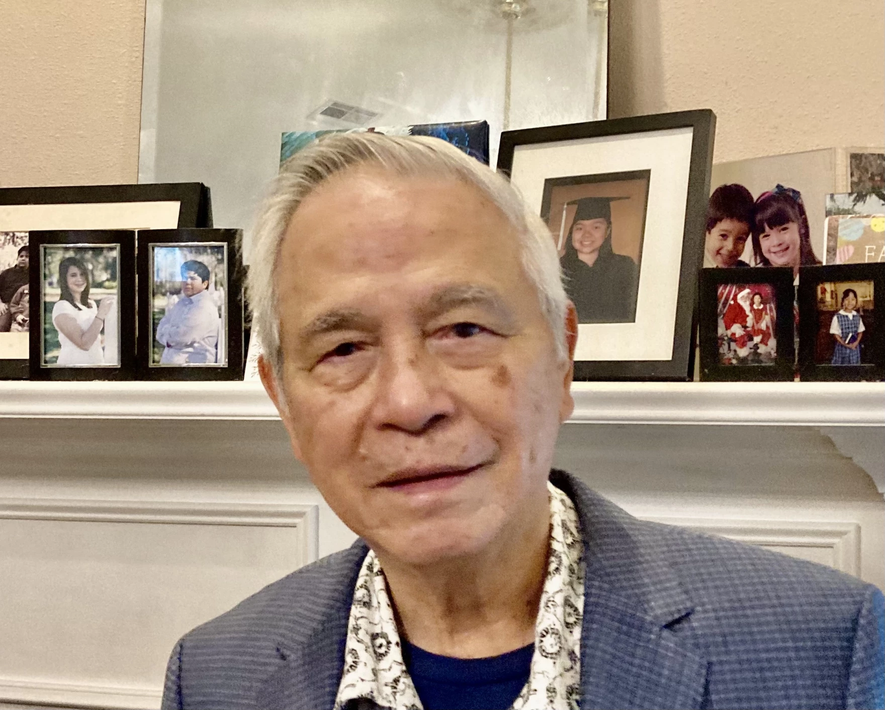 Photo of Kiem Do: an older man with gray hair and a blue blazer stands in front of a mantel displaying several framed family photos.