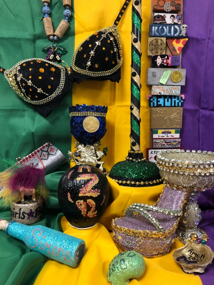 Various Mardi Gras-themed crafts are displayed, including a decorated bra, a colorful shoe, and beaded masks. The background features green, yellow, and purple fabric.