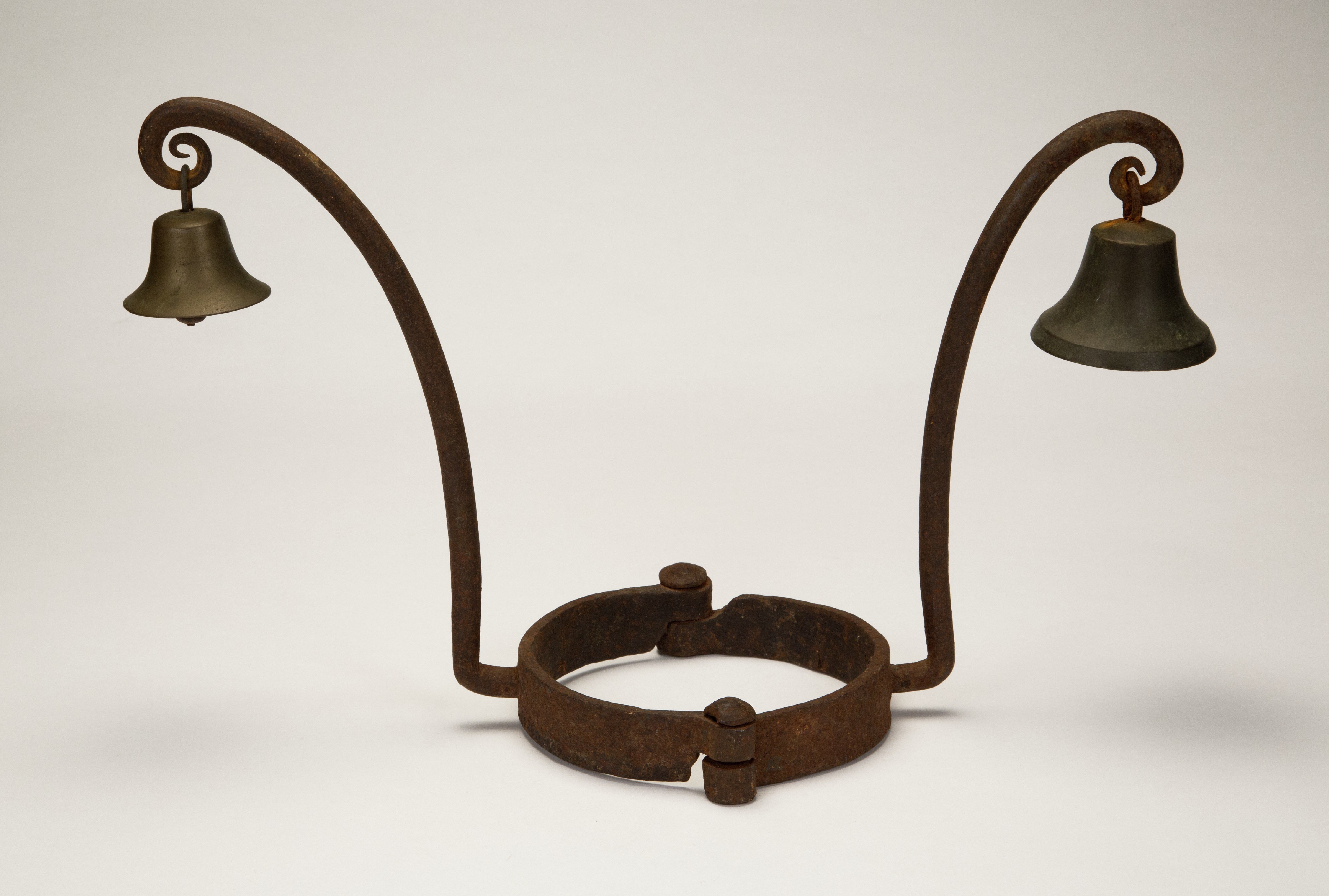 An antique, rusted metal shrews fiddle with two curved arms extending upward, each ending with a small bell. The central neck piece has a weathered appearance, indicative of its age. The background is plain and neutral.