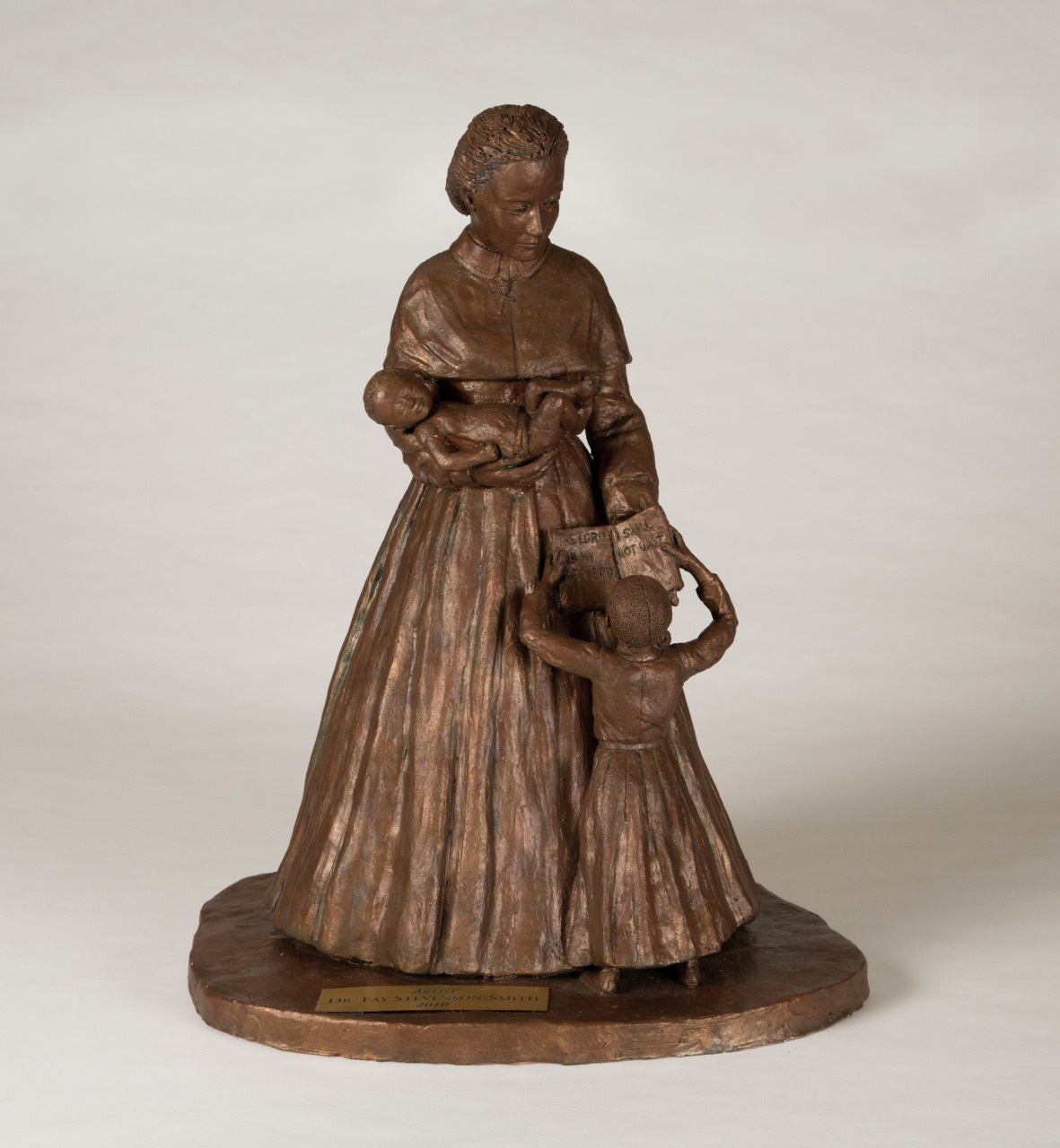 A bronze statue of a woman holding a baby and a book, while a child reaches up to touch the book. The woman wears an old-fashioned dress, and both the woman and the child stand on a circular base.