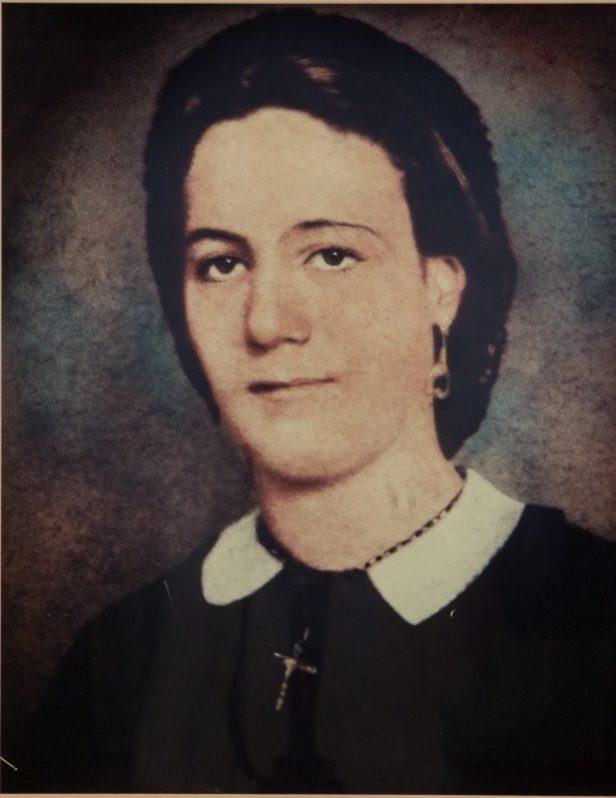 A vintage portrait of a woman with short, dark hair. She is wearing earrings, a necklace with a cross, and a dark top with a white collar. The background is softly blurred, giving the image an old-fashioned look.