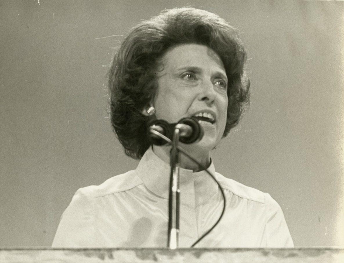 A woman with short, wavy hair and wearing a collared blouse speaks passionately at a microphone. The image has a vintage, black-and-white tone, capturing her expression and the podium in front of her.