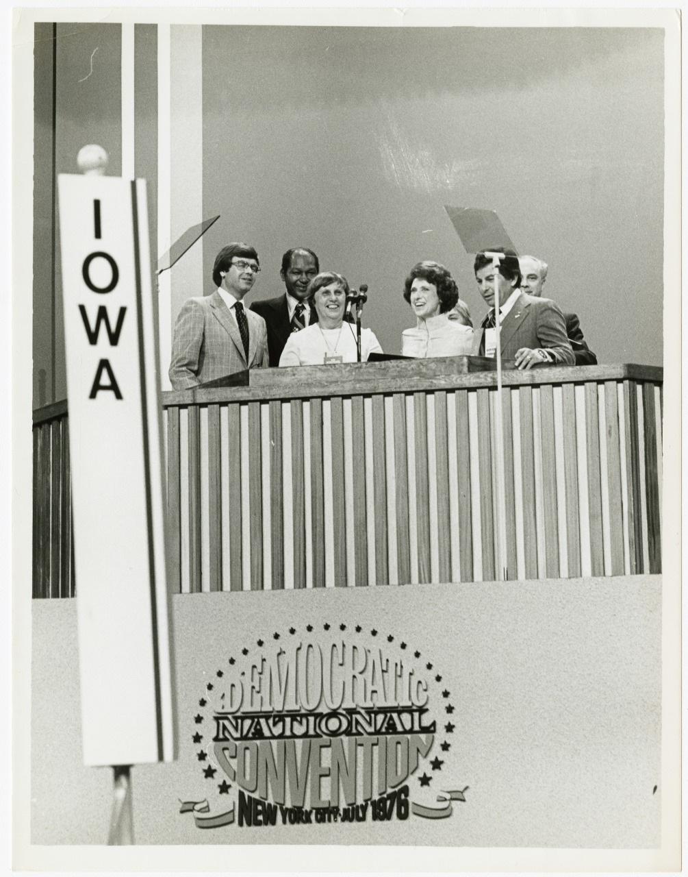 A group of people stands on a stage behind a podium at the Democratic National Convention in New York City, 1976. A sign reads IOWA in the foreground.