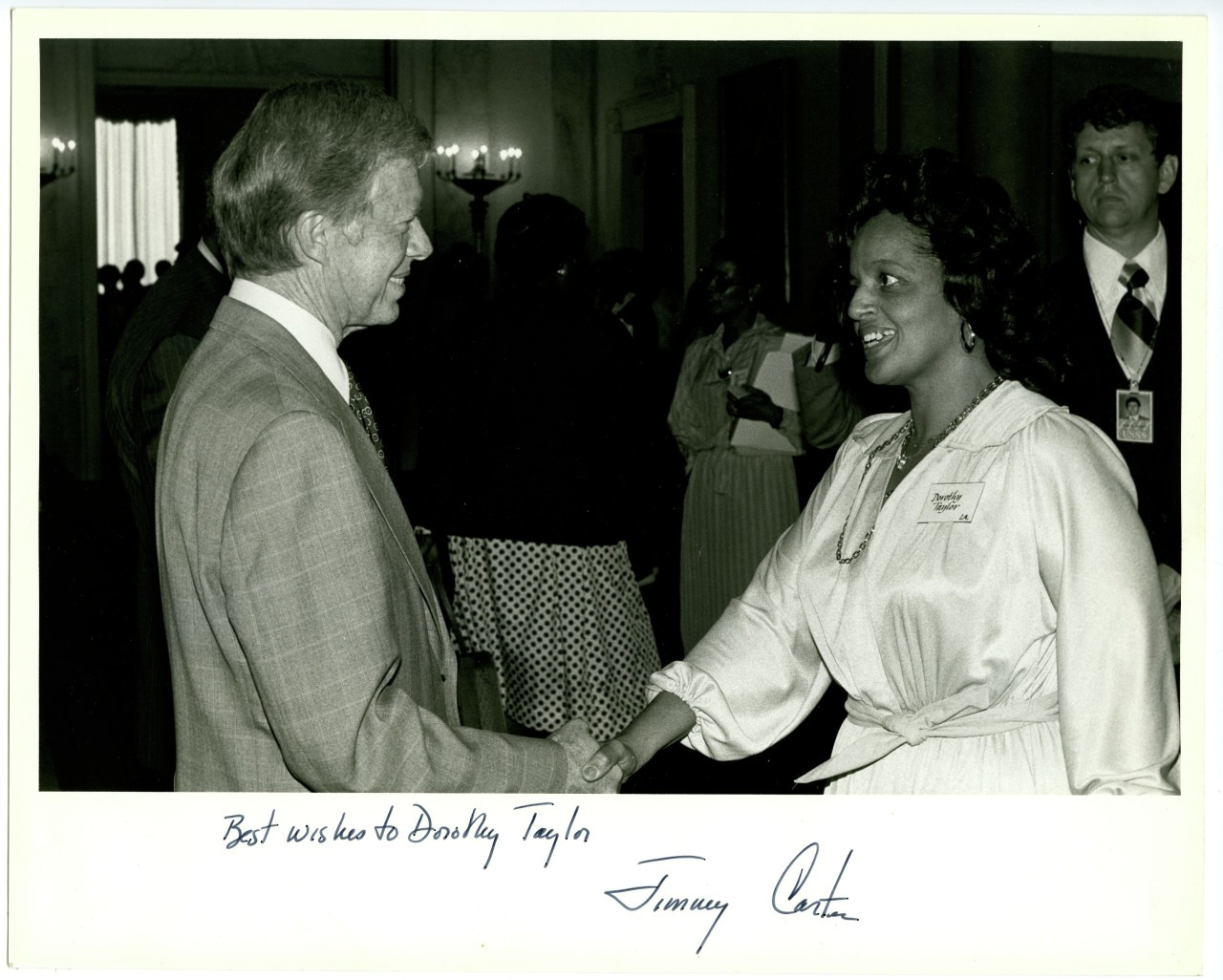 A man in a suit shakes hands with a woman in a blouse, surrounded by people in a formal setting. There is text at the bottom that reads Best wishes to Bradley Taylor signed by Jimmy Carter.