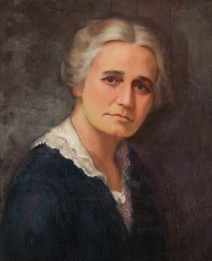 A portrait of an older woman with short, wavy gray hair. She wears a dark dress with a white lace collar and gazes softly at the viewer. The background is a muted, dark color, giving the image a classic, vintage feel.