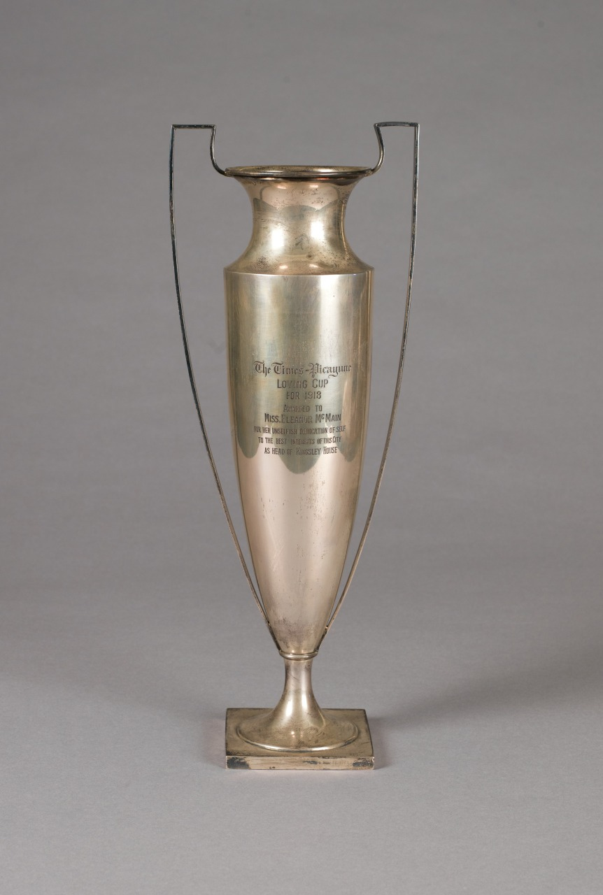 A tall, silver trophy with handles and inscriptions on its front, set against a plain gray background. The trophy has an elongated, elegant shape with a solid base.