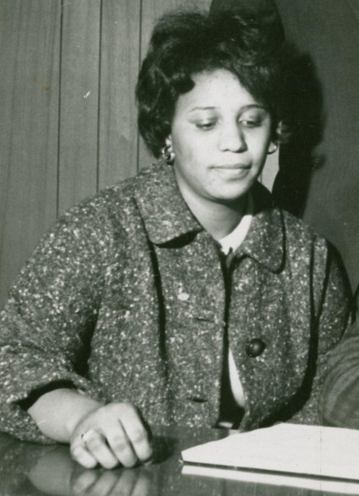 A woman with short, dark hair sits at a table, looking down at a document. She is wearing a textured coat and a white blouse. The setting appears to be indoors with wooden paneling in the background.