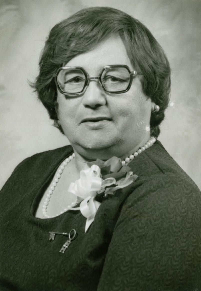 A vintage black-and-white portrait of an elderly person wearing glasses and a dark dress with a floral corsage and a pearl necklace. The individual has short hair and is facing the camera against a blurred background.