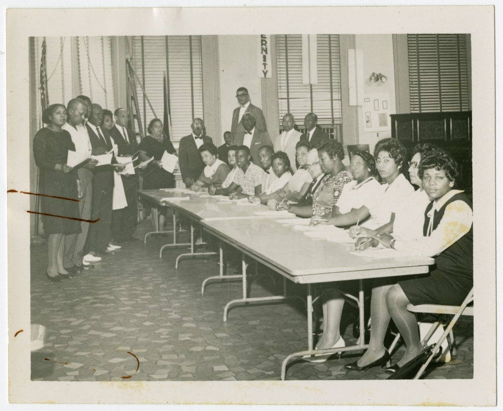 A historical black and white photo of a group of African American men and women seated and standing around a long table in a room with a tiled floor and shuttered windows. The people are dressed in formal attire.
