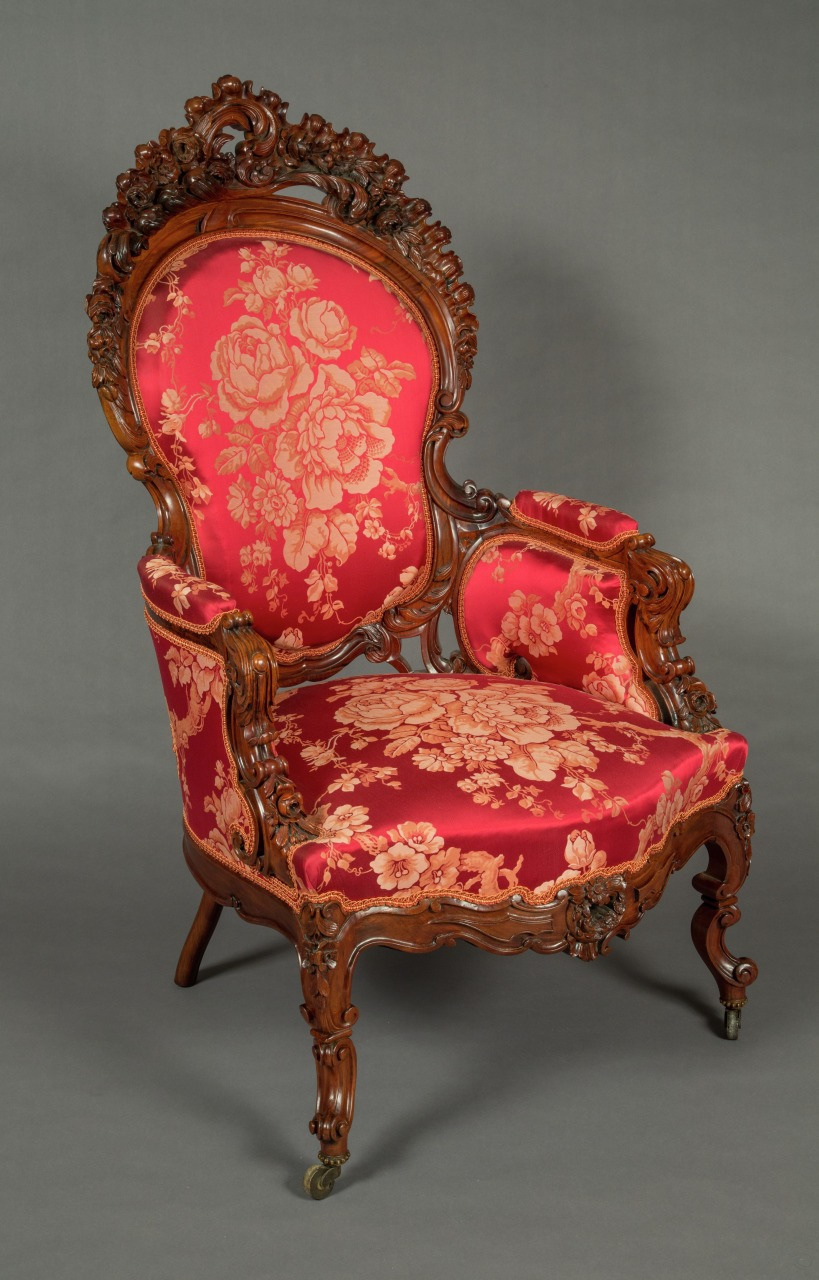 A Victorian-style armchair with ornate wooden carvings and red upholstery featuring a floral pattern. The chair has cushioned armrests and intricate detailing on the backrest and legs.