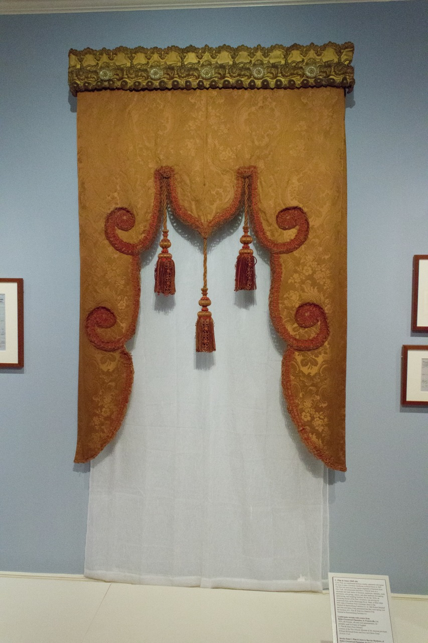 An ornate, brown and gold brocade curtain with decorative scalloped edges and red tassels hangs against a blue wall. Small framed artworks are partially visible on the sides.