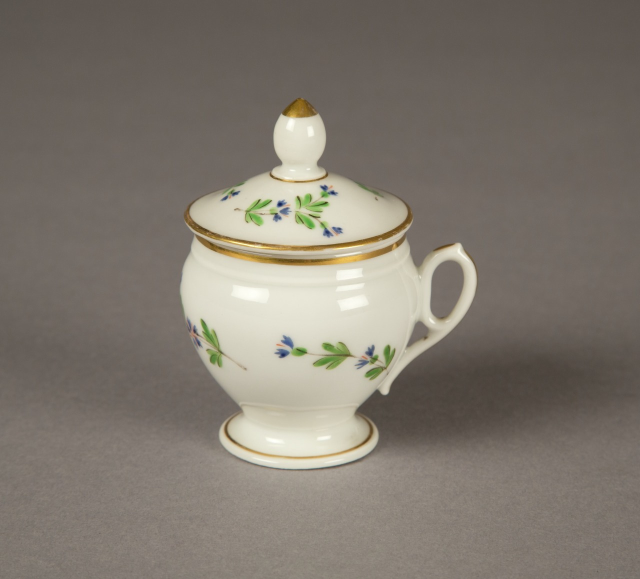 A small, white porcelain cup with a lid, adorned with blue and green floral patterns and gold trim. The cup has a delicate handle and an elegant, narrow base, resting on a smooth gray surface.