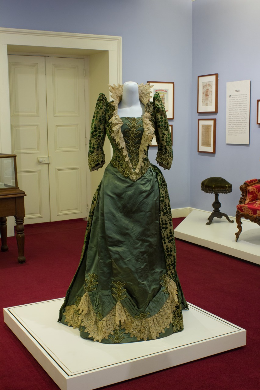 A vintage green dress with intricate lace detailing displayed in a museum setting. The dress features a fitted bodice, puffed sleeves, and an embellished skirt. The background includes framed documents and antique furniture on a burgundy carpet.