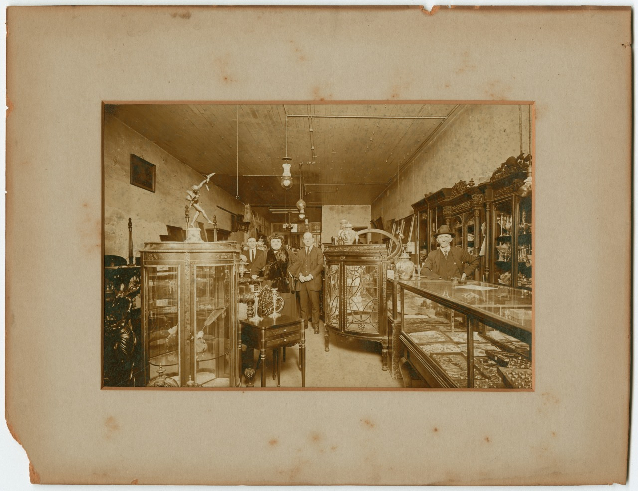 A vintage photograph of a jewelry store interior with wooden display cases and ornate decor. Three people stand behind the displays. Shelves and counters are filled with various items, and a ceiling fan is visible above.