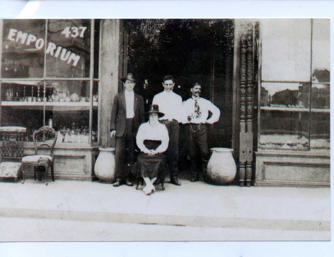 A vintage photo shows four people standing in front of a store with Emporium on the window. Two men stand on either side of a seated woman, while another man stands beside her. The storefront features ornate wooden pillars and large pots.