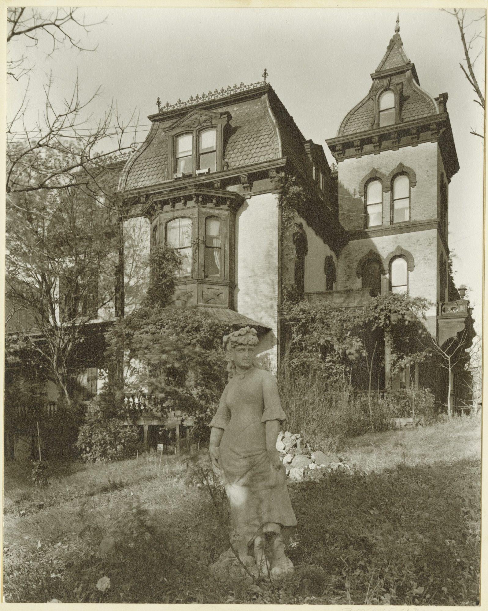 A sepia-toned photograph of a Victorian-style house with ornate details, steep roofs, and tall windows. In the foreground, there is a garden featuring a statue of a woman. The scene exudes a vintage and historical ambiance.