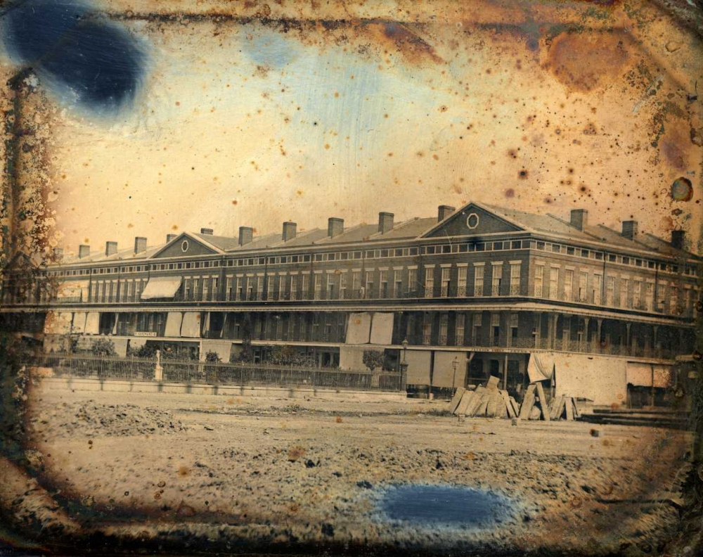 A vintage photograph shows a long, multi-story brick building with a row of windows and a pitched roof. The structure is surrounded by a dirt lot, with scattered debris in the foreground. The image is weathered and marked with spots.