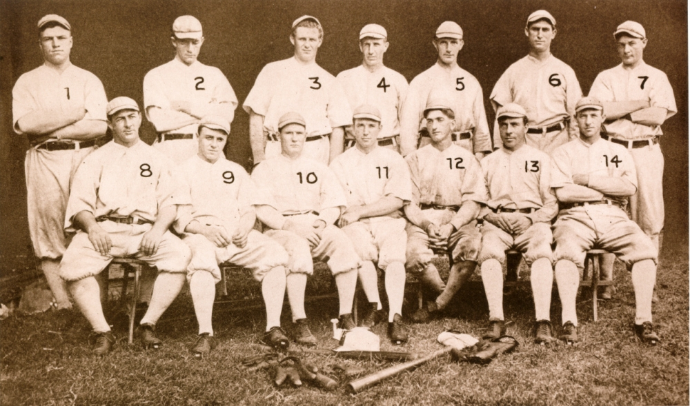 A vintage sepia photograph shows a baseball team, consisting of fourteen men in early 20th-century uniforms. A bat, gloves, and baseballs lie on the grass in front. Each player is numbered from 1 to 14.