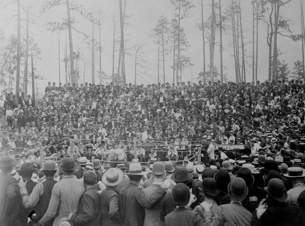 A large crowd of people in early 20th-century attire watches an outdoor boxing match. Spectators, wearing hats, fill the wooden bleachers and surround the ring. Tall trees are visible in the background.