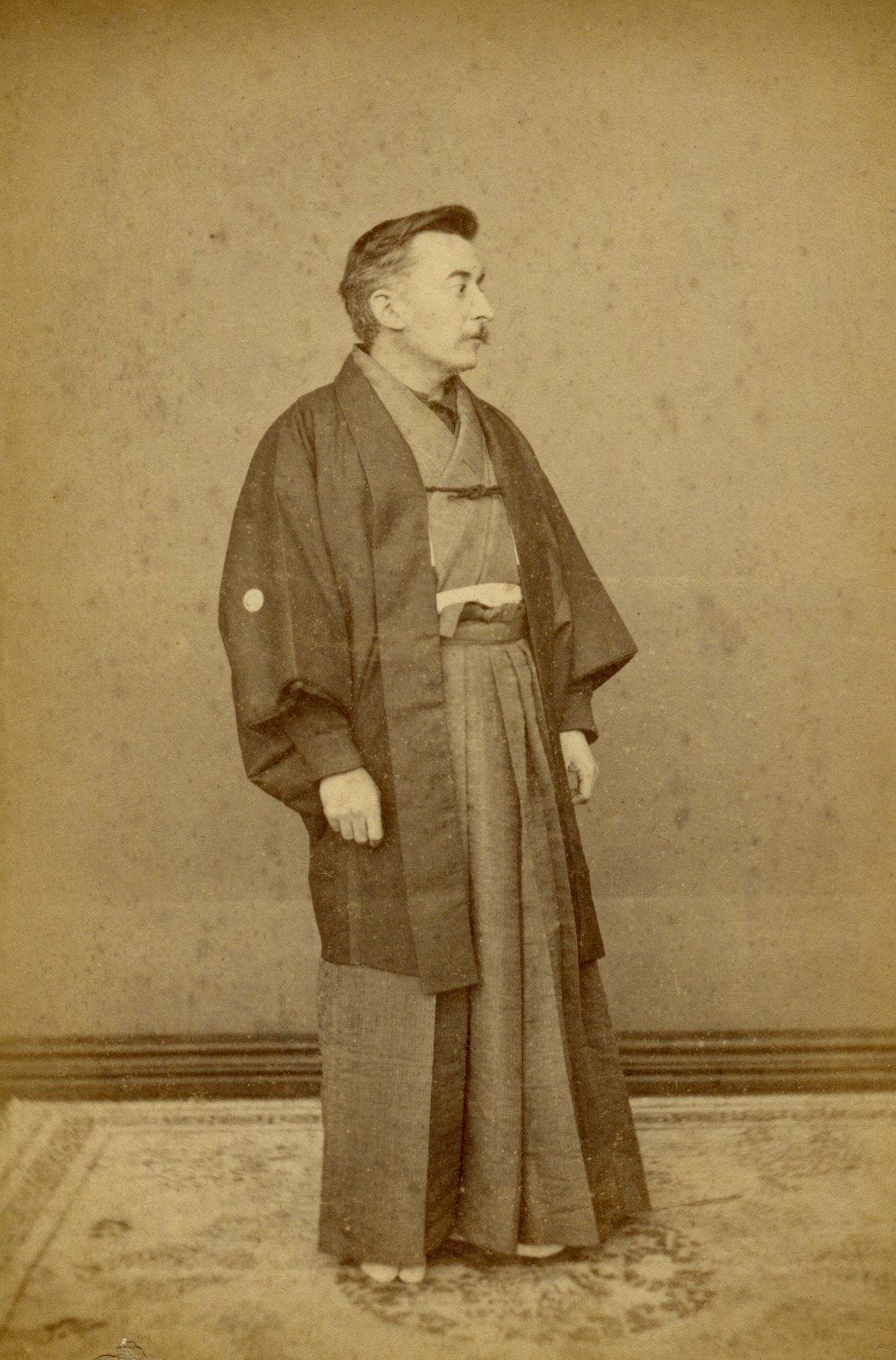 A sepia-toned photograph of a person standing in a traditional Japanese kimono and hakama, looking to the side. The floor features a patterned carpet, and the background is plain, highlighting the attire.