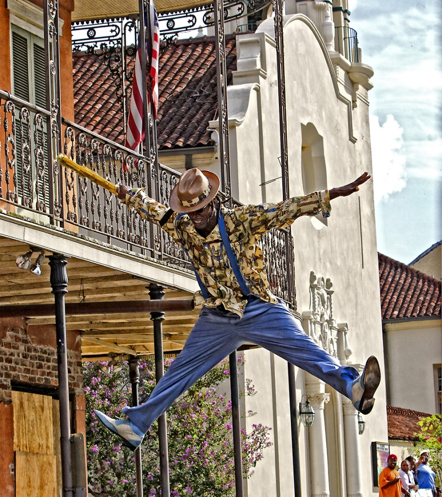 A performer in a colorful shirt and blue pants energetically jumps with arms and legs outstretched in front of a historic building. He holds a yellow stick, and American flags are displayed nearby.