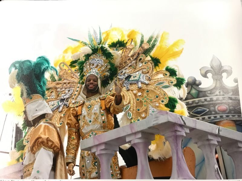 A person wearing an elaborate, colorful costume with feathers and jewels participates in a parade, standing on a float with decorative elements. The costume features gold, green, and yellow colors.