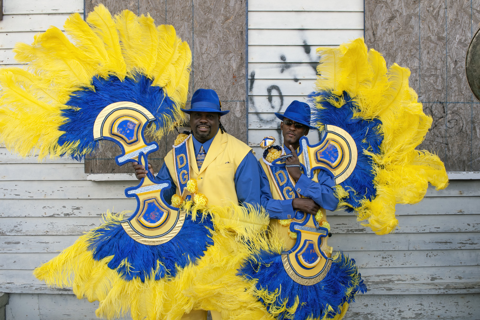 Dressed in blue and yellow suits with matching hats, two men hold ornate, feathered accessories, smiling as they pose in front of a boarded-up building.