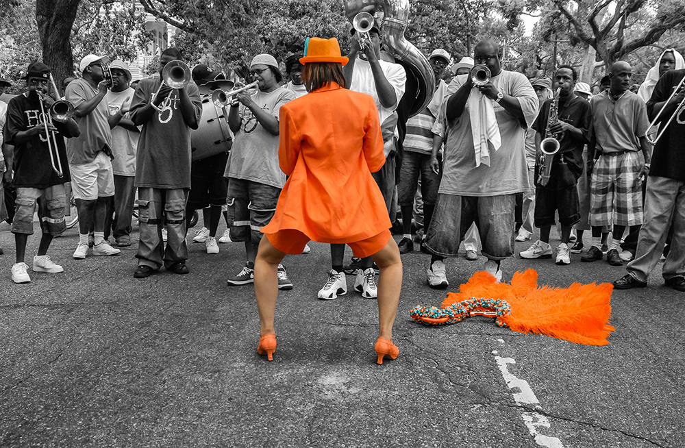 A person in an orange suit and hat energetically dances in the street facing a brass band. The band members play trumpets and trombones, while a fluffy orange prop lies on the ground next to the dancer. The background is grayscale.
