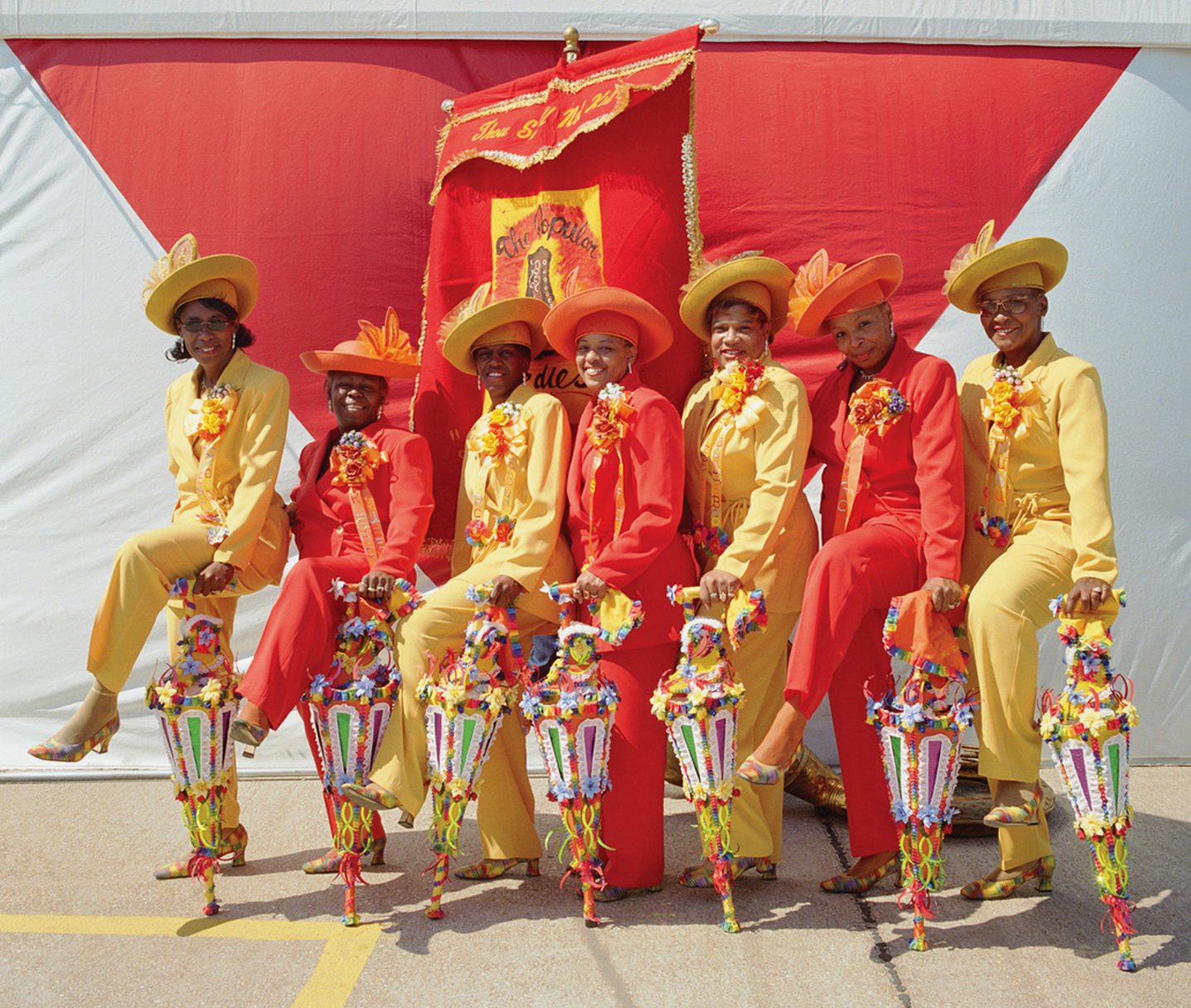 A group of eight women dressed in vibrant yellow and red suits with matching hats pose in front of a red banner, each holding a decorated parasol.