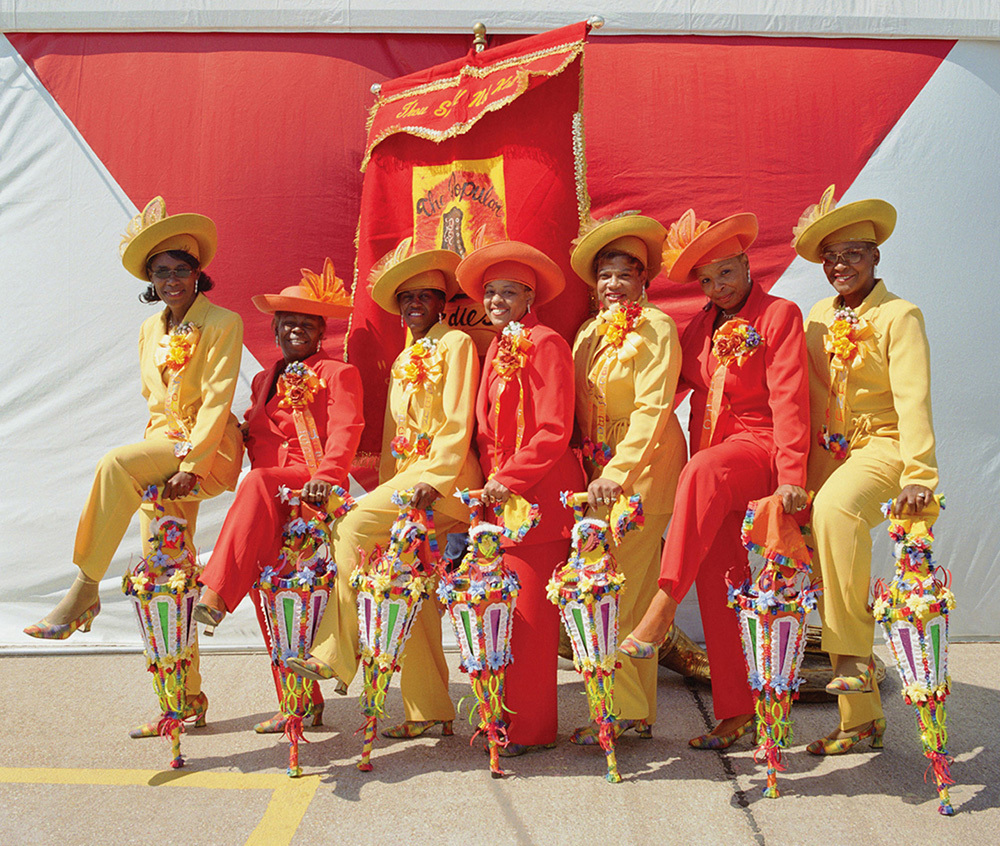 A group of eight people in coordinated red and yellow outfits and hats pose in front of a red and white backdrop. They hold colorful parasols, and a decorative red banner is displayed behind them.