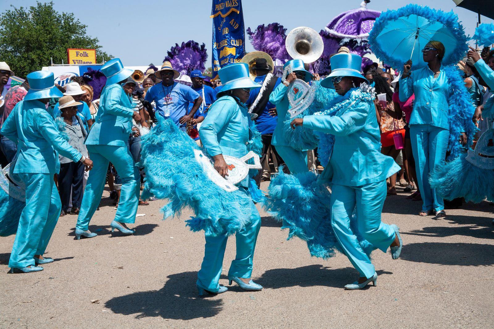 A vibrant parade with participants dressed in matching bright blue suits, hats, and feathered accessories. They are energetically dancing in the street, surrounded by a crowd. Some carry blue parasols, and musicians play instruments in the background.