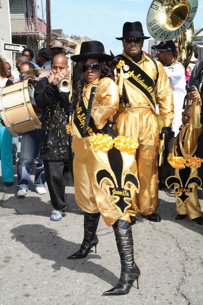 A group of musicians and dancers, dressed in gold costumes and black hats, parade with instruments, including a trumpet and sousaphone, on a city street. The clothing features fleur-de-lis designs, and a woman poses confidently in front.