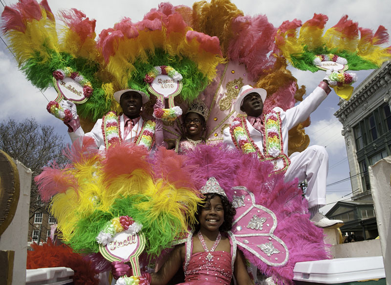 A colorful Mardi Gras parade scene featuring four people in vibrant feathered costumes and wide-brimmed hats in pink, green, and yellow. A child dressed in pink sits in front, smiling.