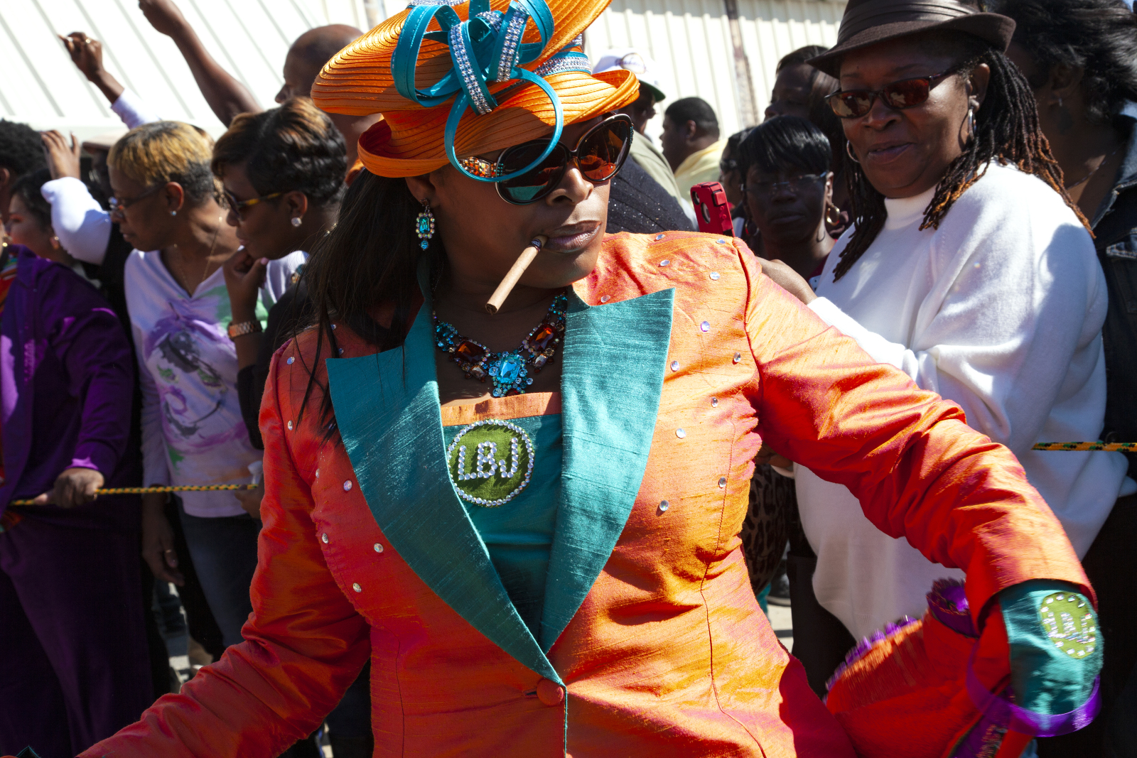 A woman in a bold orange and teal outfit dances confidently in a parade, wearing a matching hat, sunglasses, and jewelry while holding a cigar in her mouth. A crowd watches in the background.