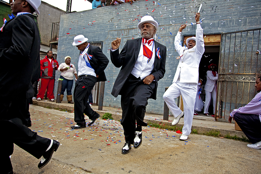 A group of men dressed in sharp black and white suits with red, white, and blue accents energetically dance in the street as confetti rains down. An older man in the center, wearing a white hat and two-tone shoes, leads the procession with graceful footwork. A crowd watches from the sidelines and a balcony, while a man in white jumps with excitement near an open doorway.