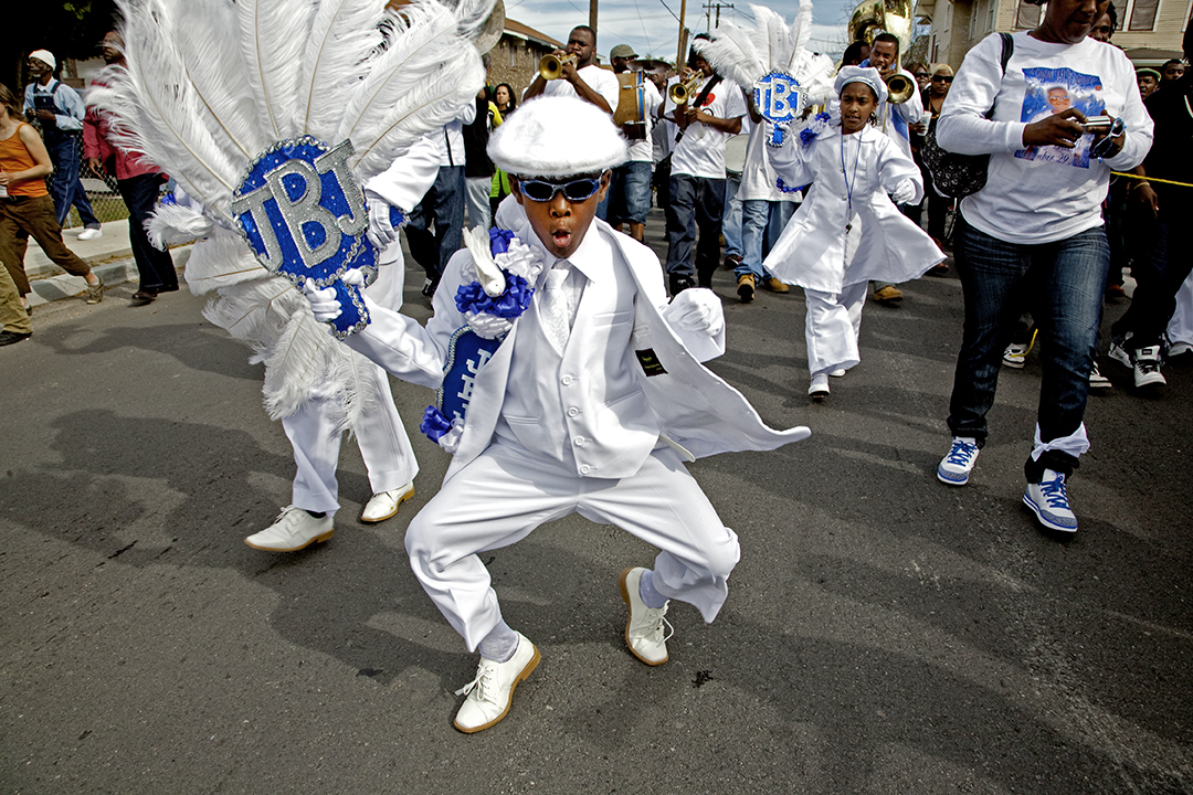 A child in a white suit, feathered accessories, and sunglasses performs energetically in the street, holding a decorated sign. Other parade participants, including musicians, follow behind.